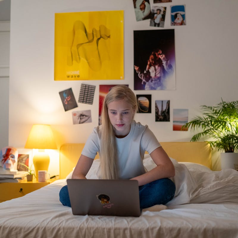 Teen girl using a laptop in her cozy bedroom lit with a warm glow, perfect for remote work or study.